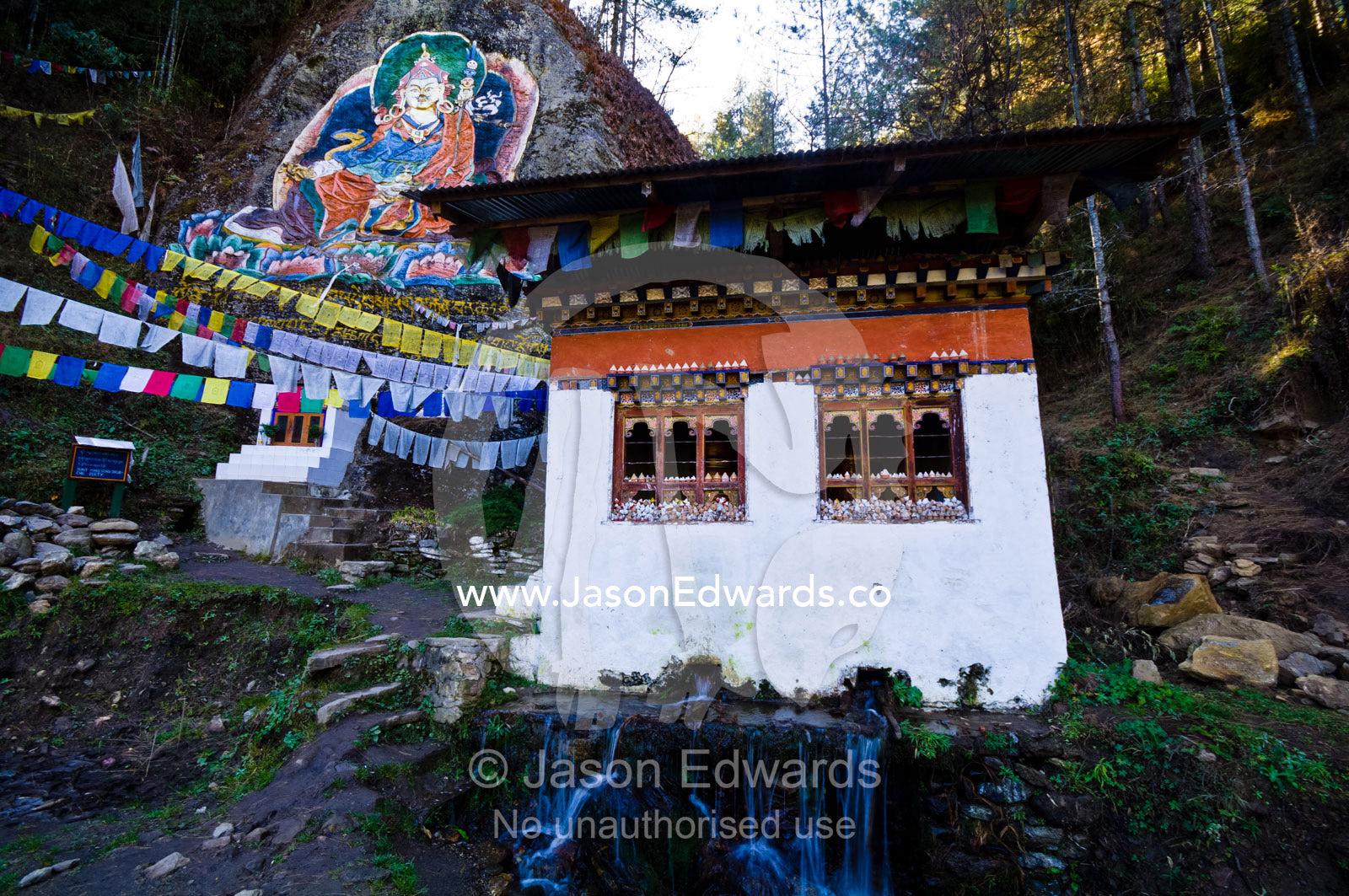 A Mani Chukhor built over a small stream is overlooked by a mural of Guru Rinpoche. Thimphu District, Bhutan.