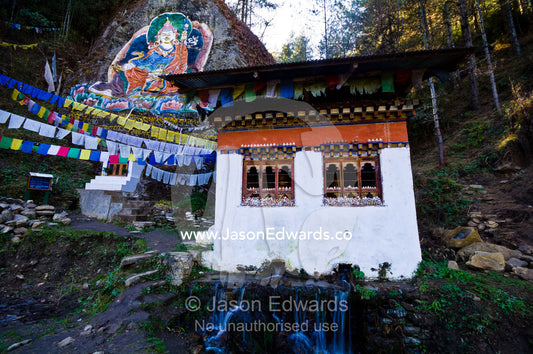 A Mani Chukhor built over a small stream is overlooked by a mural of Guru Rinpoche. Thimphu District, Bhutan.