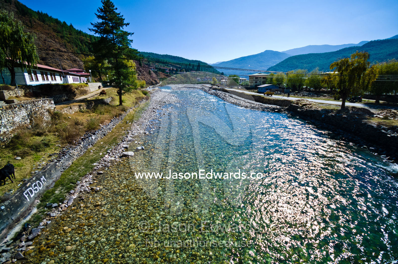 The clear turquoise alpine waters of the Paro River travel past a Buddhist monastery. Paro Chu, Paro River, Rinpung Dzong, Paro Dzong, Paro, Paro District, Bhutan.