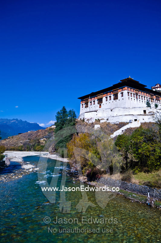 The clear turquoise alpine waters of the Paro River travel past a Buddhist monastery. Paro Chu, Paro River, Rinpung Dzong, Paro Dzong, Paro, Paro District, Bhutan.