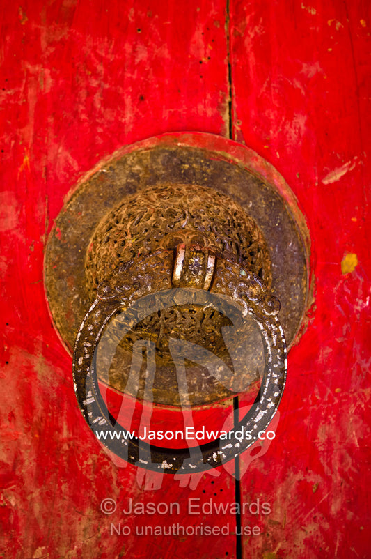 A rusting door knocker on a weathered bright red door leading to a Buddhist monastery. Gangteng Monastery, Ganteng, Wangdue Phodrang District, Phobjika Valley, Phobjikha Valley, Gangteng Valley, Black Mountains, Bhutan.