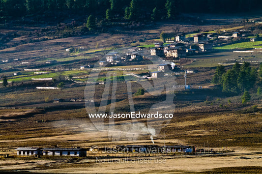 Smoke from a kitchen in a farmhouse floats into the air above a village overlooking a Himalaya wetland. Phobjikha Conservation Area, Phobjika Valley, Phobjikha Valley, Gangteng Valley, Black Mountains, Bhutan.