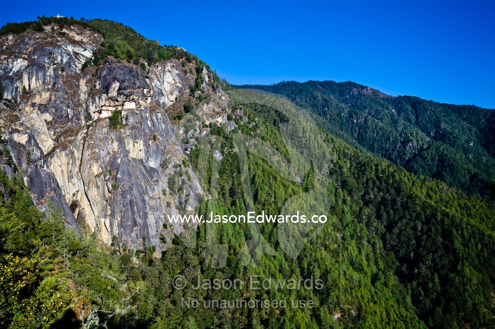 An ancient Buddhist monastery perched on a sheer cliff face above a forested valley in the Himalaya. Paro Taktsang, The Tigers Nest, Paro Valley, Paro District, Bhutan.