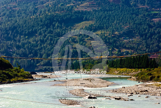 A long suspension bridge spans a wide alpine river flowing through a Himalaya valley. Paro River, Paro Chu, Paro, Paro District, Bhutan.