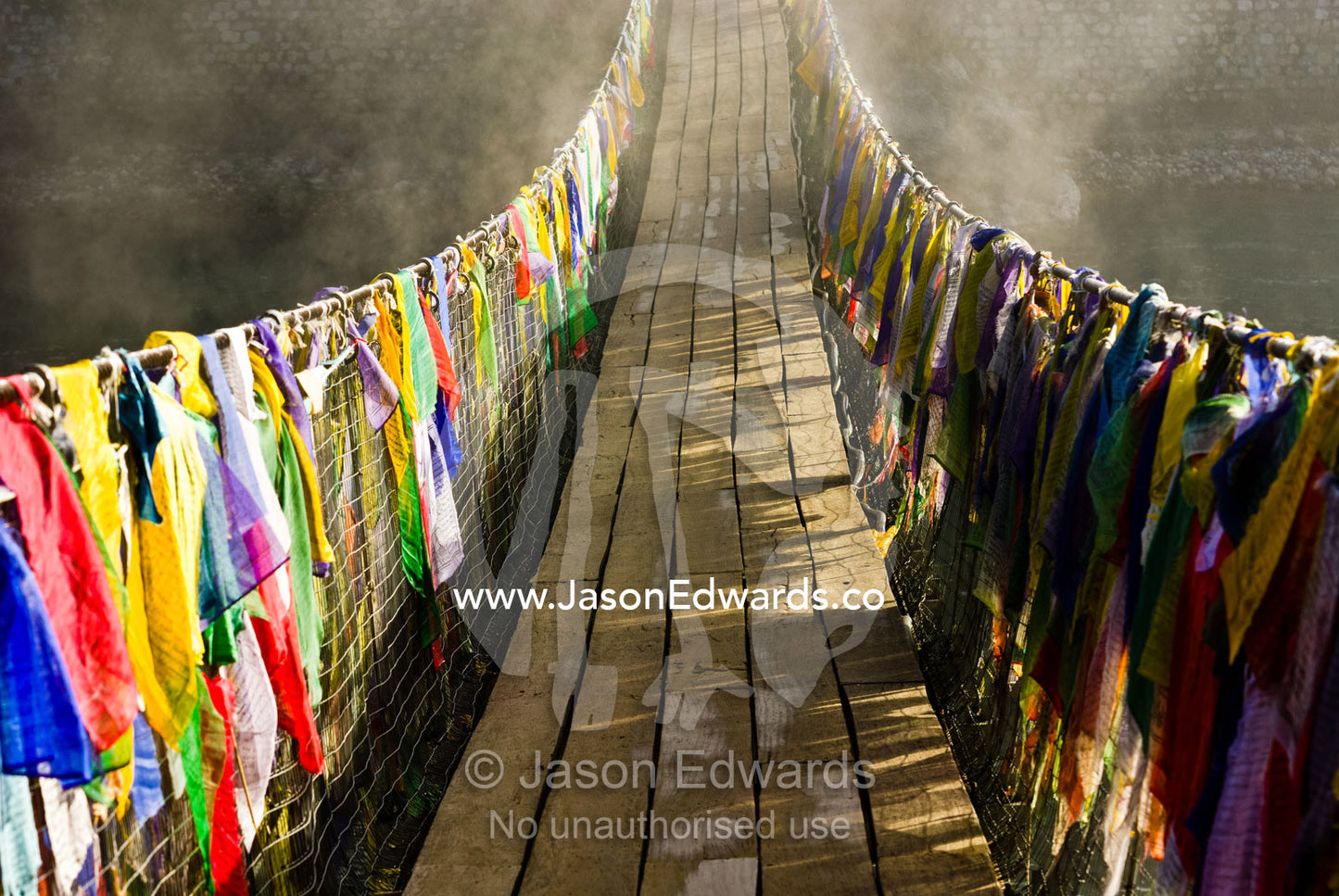 Prayer flags drape a suspension bridge across a river as dawn melts the fog away. Punakha River, Punakha Valley, Punakha-Wangdiphodrang District, Bhutan.