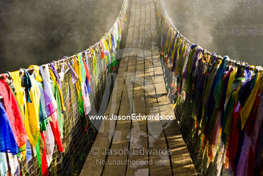 Prayer flags drape a suspension bridge across a river as dawn melts the fog away. Punakha River, Punakha Valley, Punakha-Wangdiphodrang District, Bhutan.
