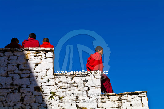Buddhist monks in bright red robes resting in the sunshine on the ancient white-washed stone steps of a monastery. Rinpung Dzong, Paro Dzong, Paro, Paro District, Bhutan.