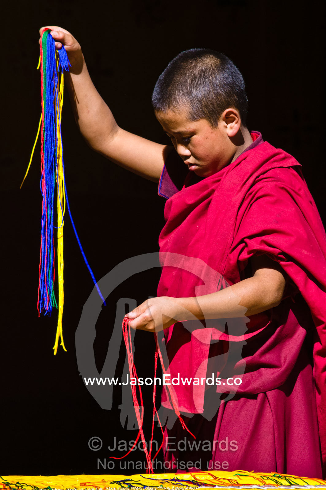 A young Buddhist monk in red robes separates colored strips of wool to weave together. Rinpung Dzong, Paro Dzong, Paro, Paro District, Bhutan.