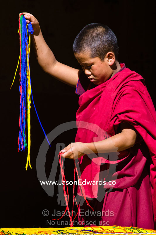 A young Buddhist monk in red robes separates colored strips of wool to weave together. Rinpung Dzong, Paro Dzong, Paro, Paro District, Bhutan.