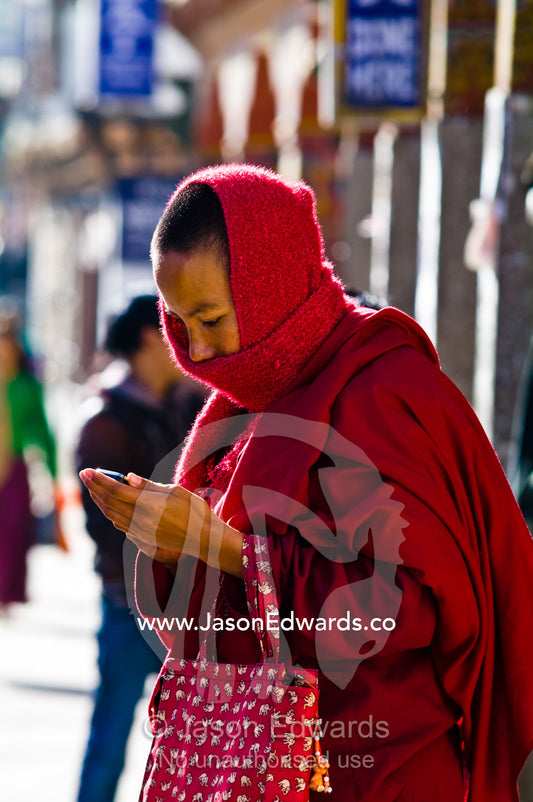 A Buddhist monk in red robes texting on a cell phone in the business district of a Himalayan town. Thimphu, Thimphu District, Bhutan.