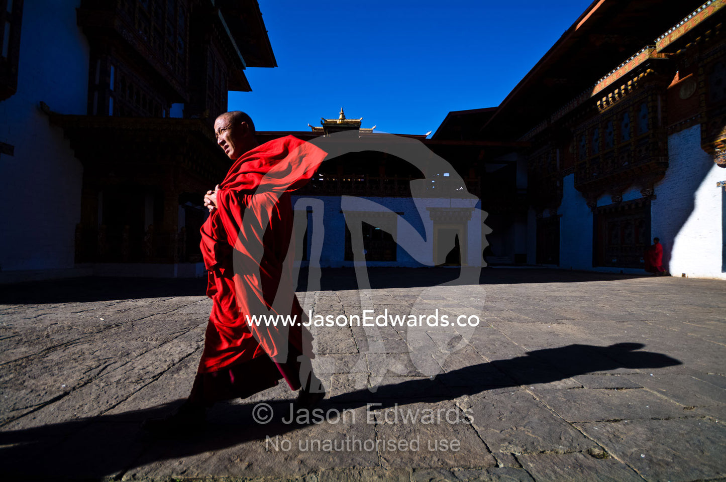 A Buddhist monk strolls across the paved courtyard of an ancient monastery with his red robes blowing in the wind. Punakha Dzong, Punakha dzongkhag, Punakha District, Bhutan.