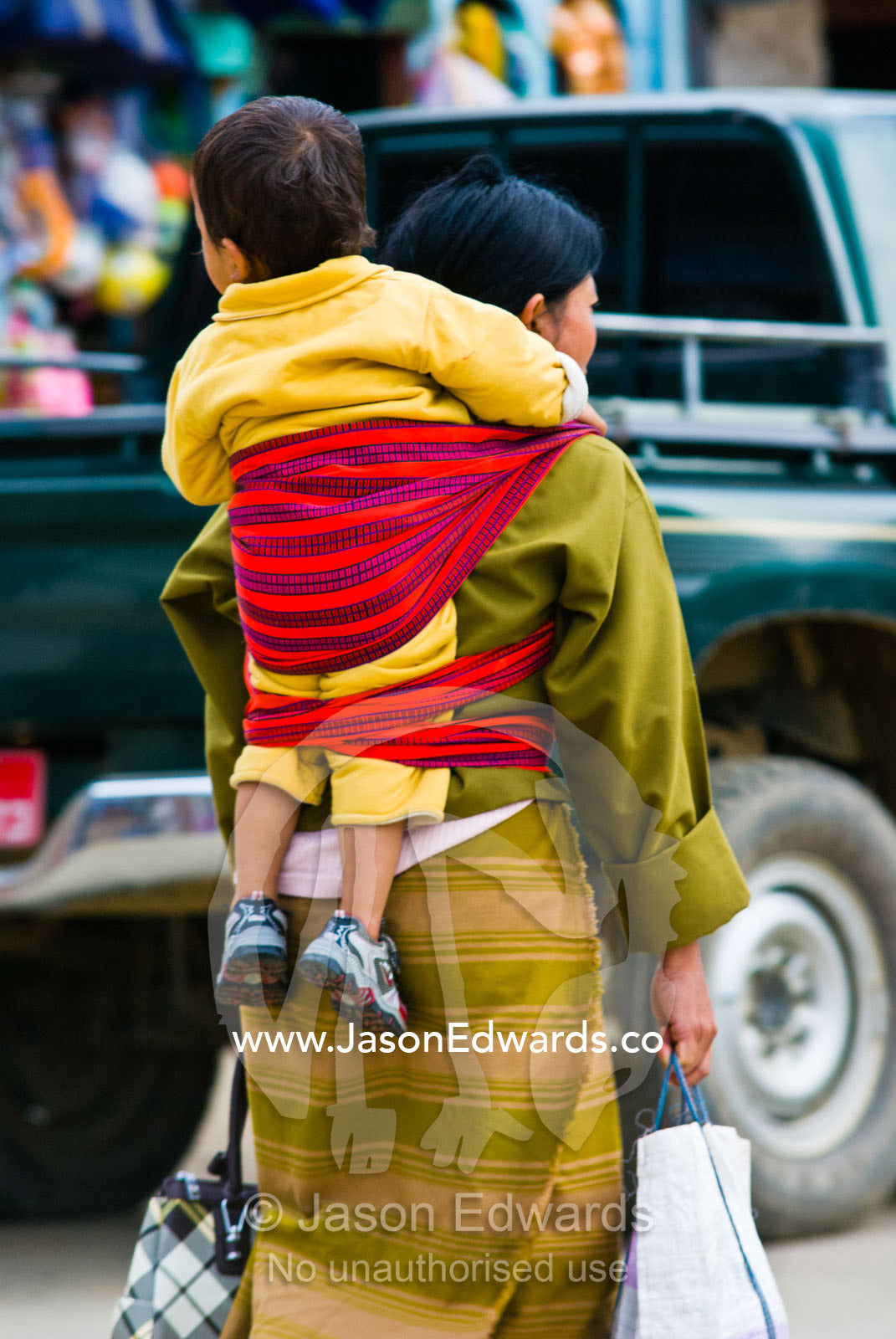 A mother carrying her baby on her back through the central business district of a Himalayan town. Wangdi, Wangdue Phodrang, Wangdue Phodrang District, Bhutan.
