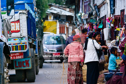 Women shopping in a roadside store whilst construction trucks and traffic congestion fill the main street. Wangdi, Wangdue Phodrang, Wangdue Phodrang District, Bhutan.