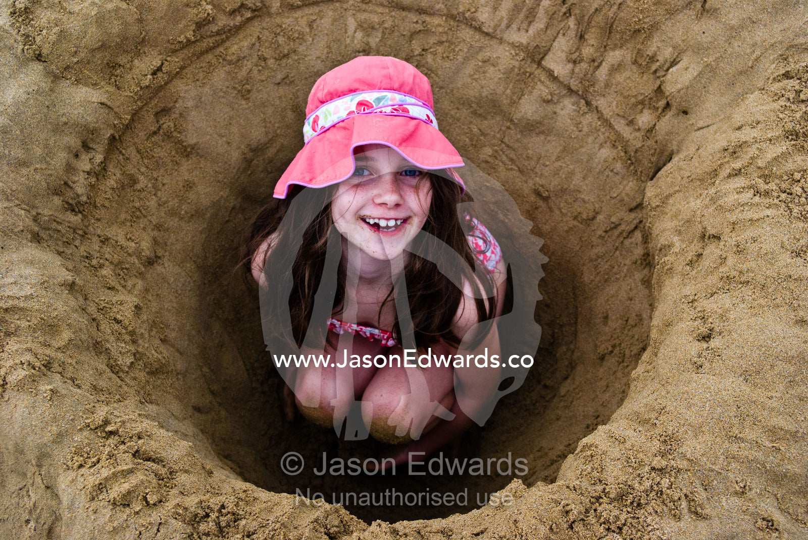 A girl in a sun hat digs a deep hole on a beach and hides inside it. Torquay, Surf Coast, Victoria, Australia.