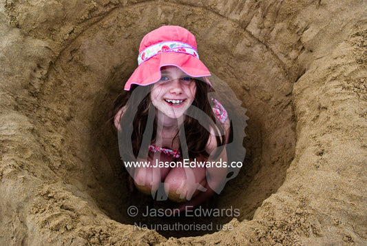 A girl in a sun hat digs a deep hole on a beach and hides inside it. Torquay, Surf Coast, Victoria, Australia.