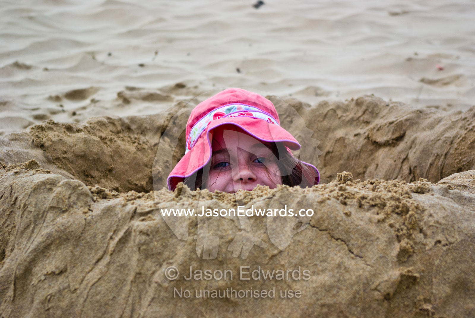 A girl in a sun hat digs a deep hole on a beach and hides inside it. Model release on file for Imogen Jackson and William Edwards.