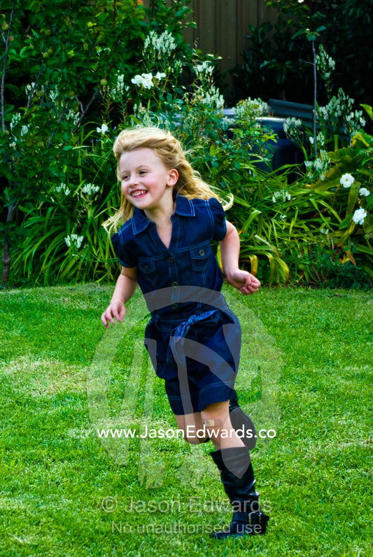 A young girl in a denim dress running and playing in a garden yard. Malvern East, Melbourne, Victoria, Australia.