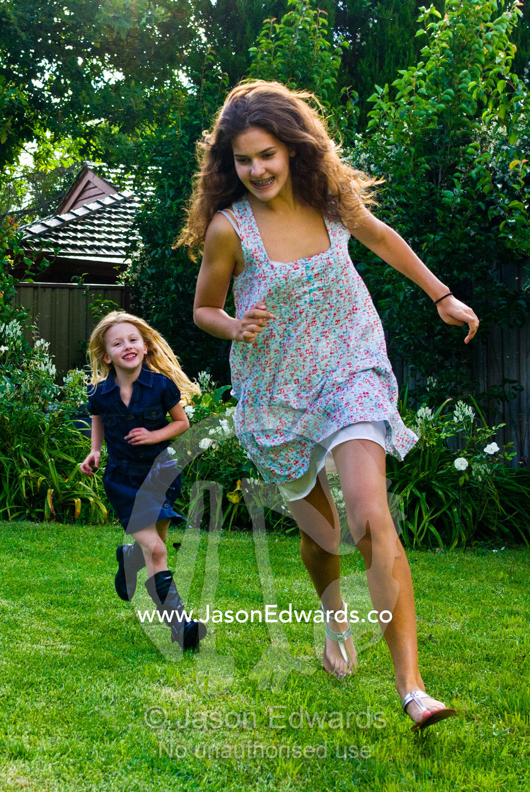 A young girl chases a teenage girl running and playing in a backyard. Malvern East, Melbourne, Victoria, Australia.