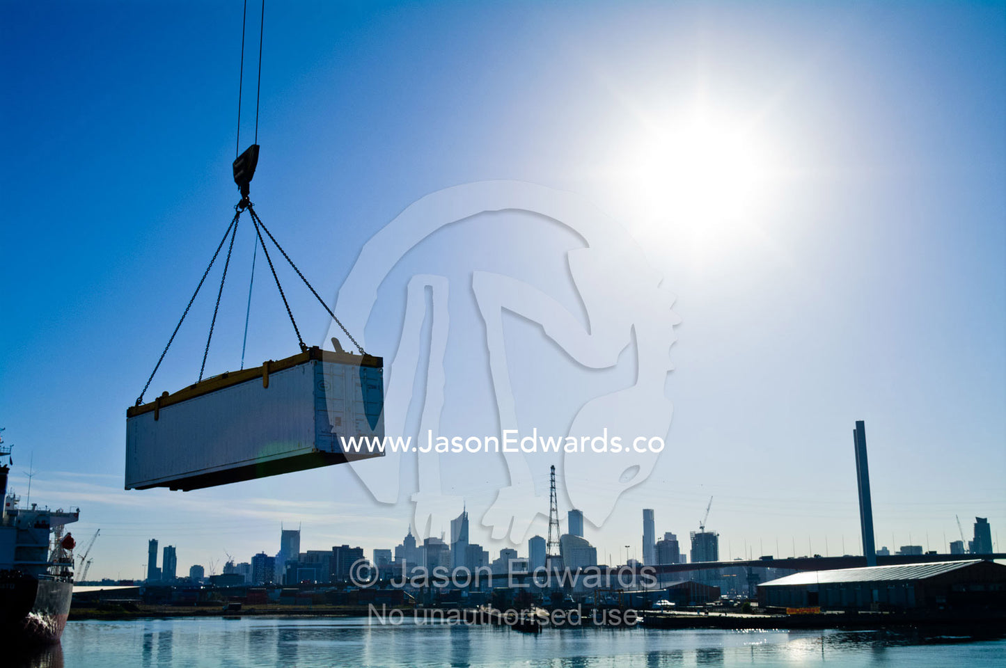 A crane lifts a shipping container over a river past a city skyline. Yarra River, Appleton Dock, Port of Melbourne, West Melbourne, Victoria, Australia.