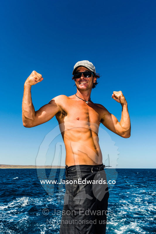 A tanned fit man flexes his bicep muscles on a boat at sea. Exmouth, Ningaloo Reef, Ningaloo Marine Park, World Heritage Site, Western Australia, Australia.