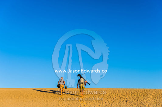 A cameraman and sound recordist hike a desert sand dune with tripod. Cape Range National Park, Exmouth, Ningaloo Reef, Western Australia, Australia.