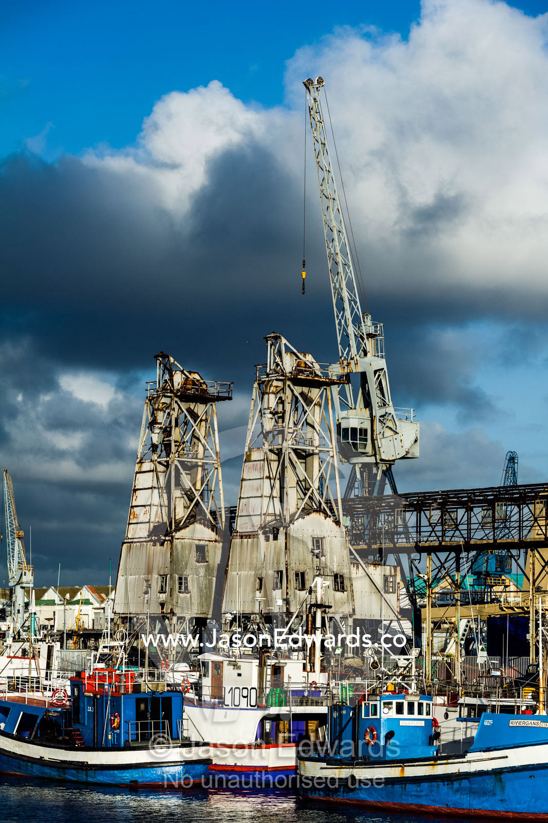 Cranes and factories line a wharf where fishing boats are anchored. Cape Town, Victoria and Alfred Waterfront, Western Cape, South Africa.