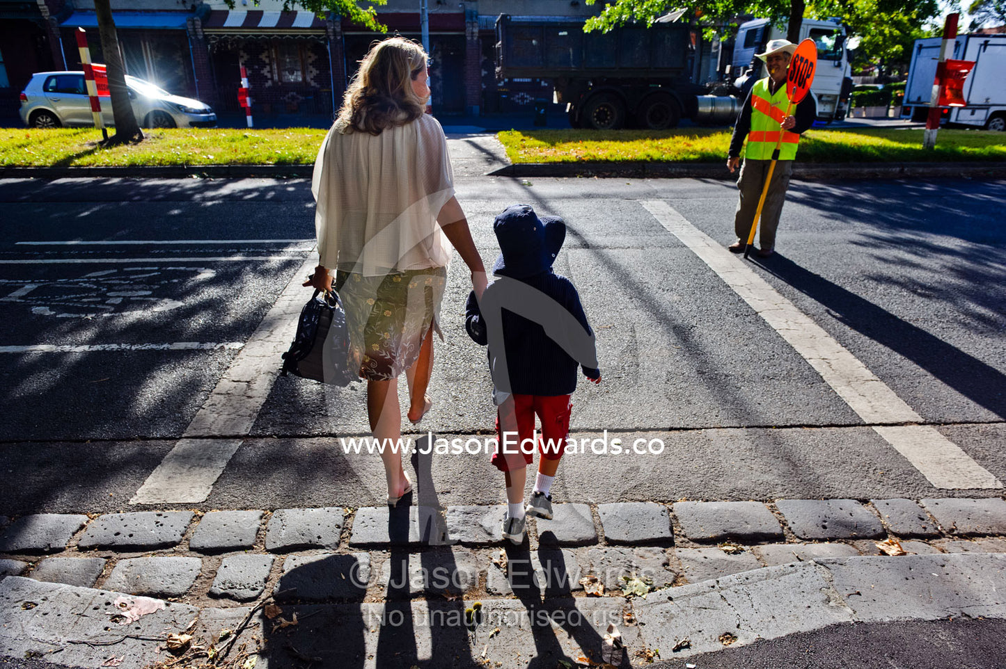 A mother crosses a busy road with her son on his first day of school. Melbourne, Carlton North, Victoria, Australia.