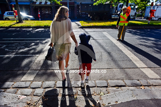 A mother crosses a busy road with her son on his first day of school. Melbourne, Carlton North, Victoria, Australia.