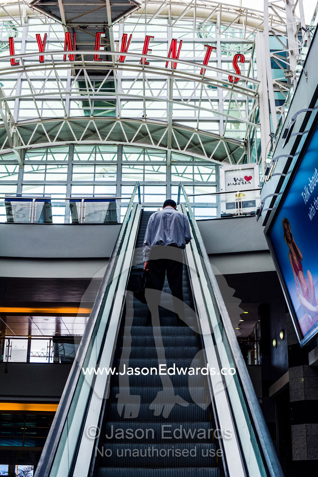 A man rides an escalator to an upper level in a shopping mall. Menlyn Park, Pretoria, Menlyn Mall, Pretoria, Gauteng Province, South Africa.