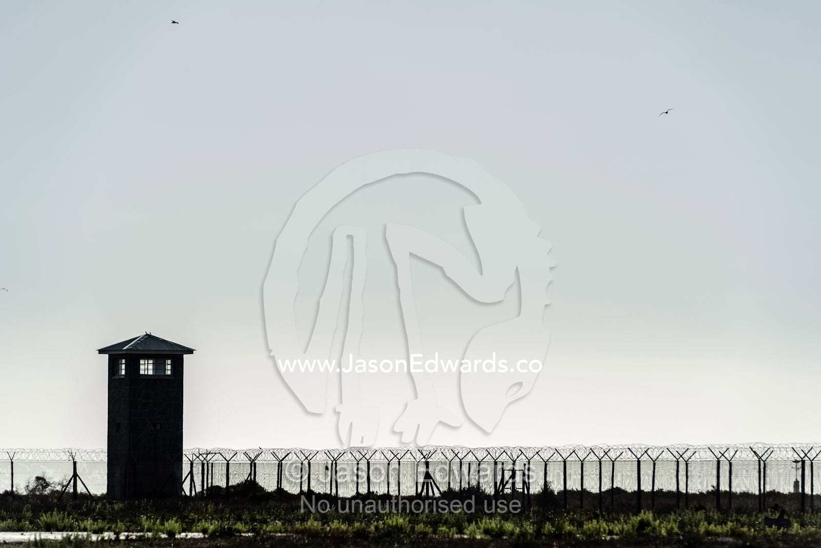 Rows of steel and wire mesh fencing surround a prison and guard tower. Robben Island, Cape Town, Bloubergstrand, Table Bay, Western Cape, South Africa.