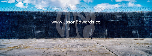 A barren featureless concrete prison yard wall topped with razor wire. Robben Island, Cape Town, Bloubergstrand, Table Bay, Western Cape, South Africa.
