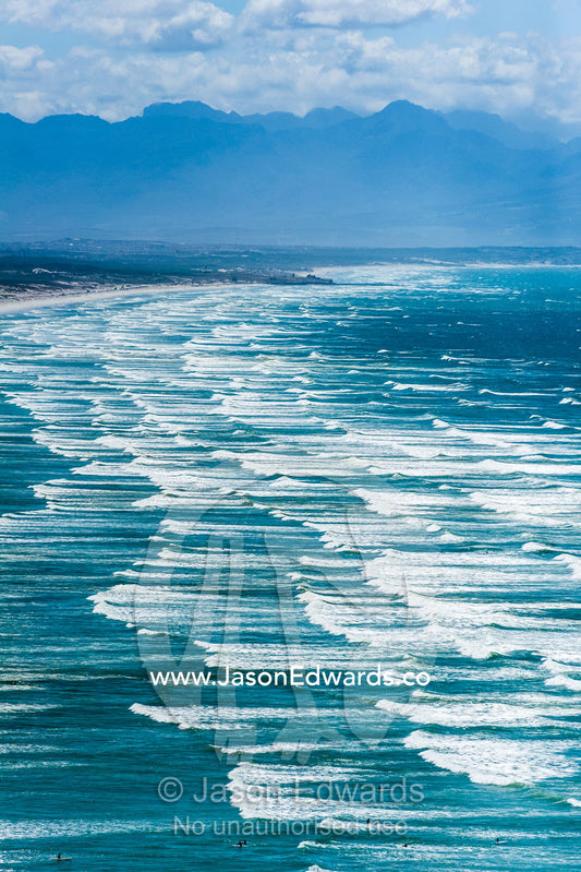 Endless waves roll onto a long, white sandy beach under a summer sky. Surfers Corner, Muizenberg, Cape Town, Chapmans Peak Drive, Western Cape, South Africa.