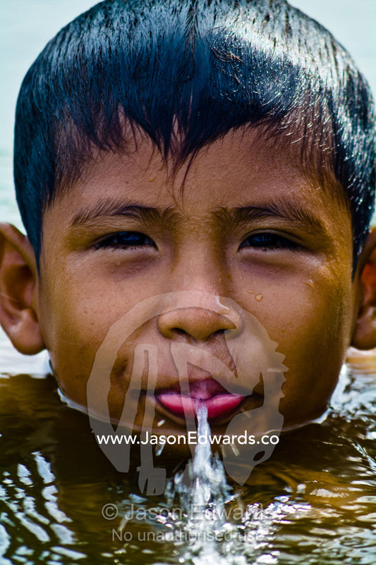 A boy swimming and squirting water from his mouth in a lake on a hot weekend afternoon. Quistococha Lake, Lago de Quistococha, Parque de Quistococha, Iquitos, Amazon Basin, Loreto Region, Maynas Province, Peru.