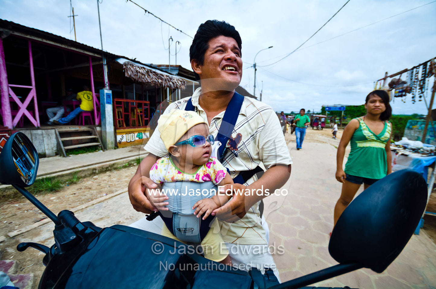 A father carries his baby wearing sunglasses in a papoose on a motorbike in a river town. Iquitos, Loreto Region, Maynas Province, Amazon Basin, Peru.
