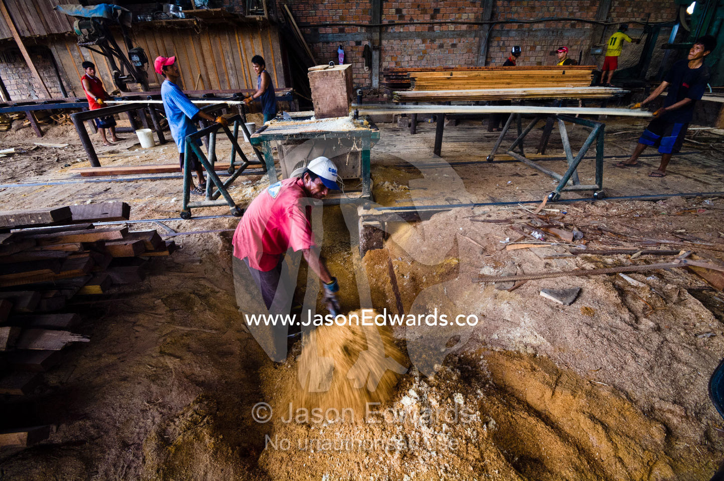 A laborer in a logging mill shovels sawdust from underneath a jigsaw blade. Puerto Masusa, Iquitos, Amazon Basin, Loreto Region, Maynas Province, Peru.