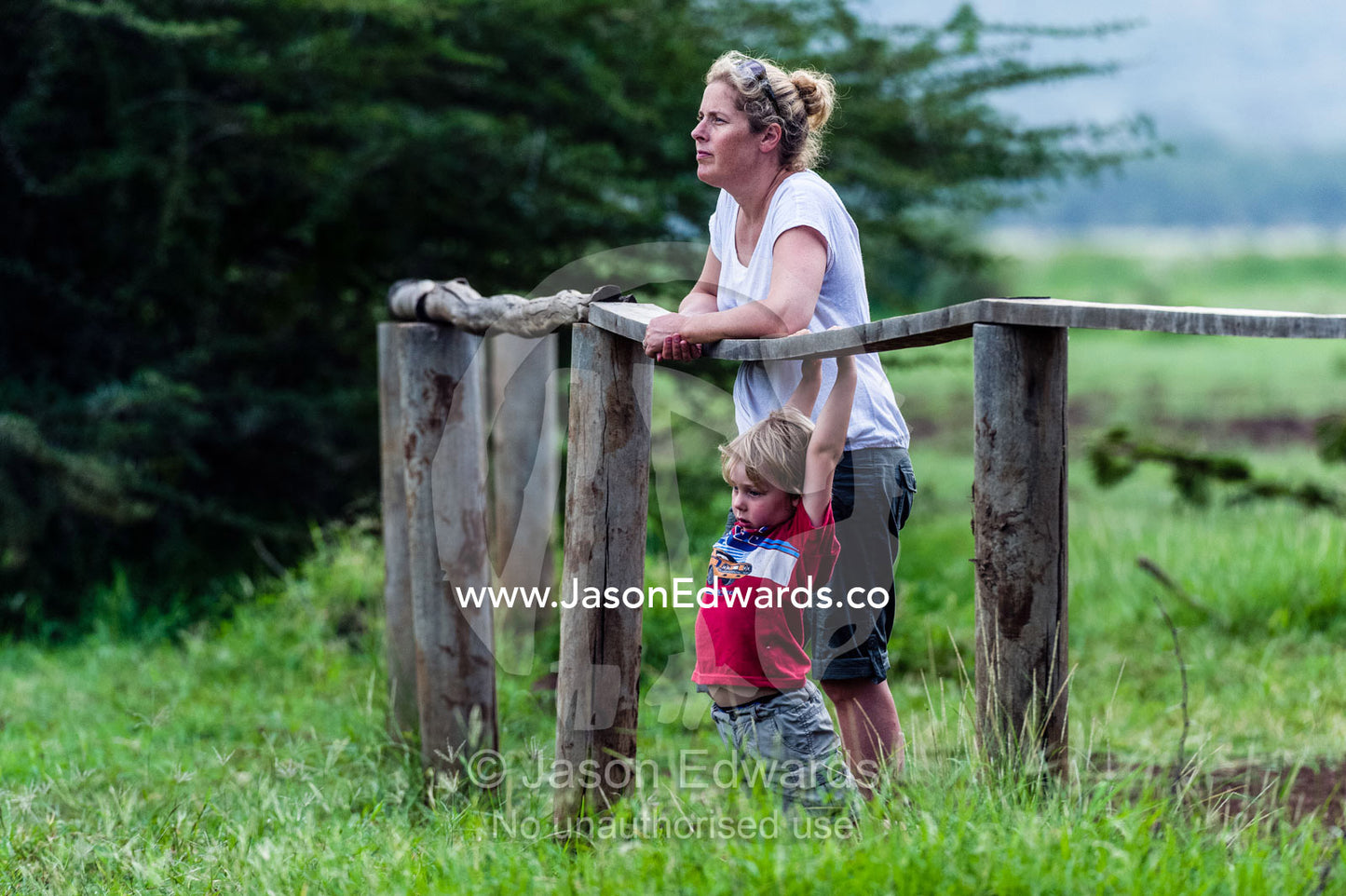 A bored boy swings on a fence rail whilst his mother watches hippos on safari. Lake Manyara National Park, Arusha Region, Tanzania, Africa.