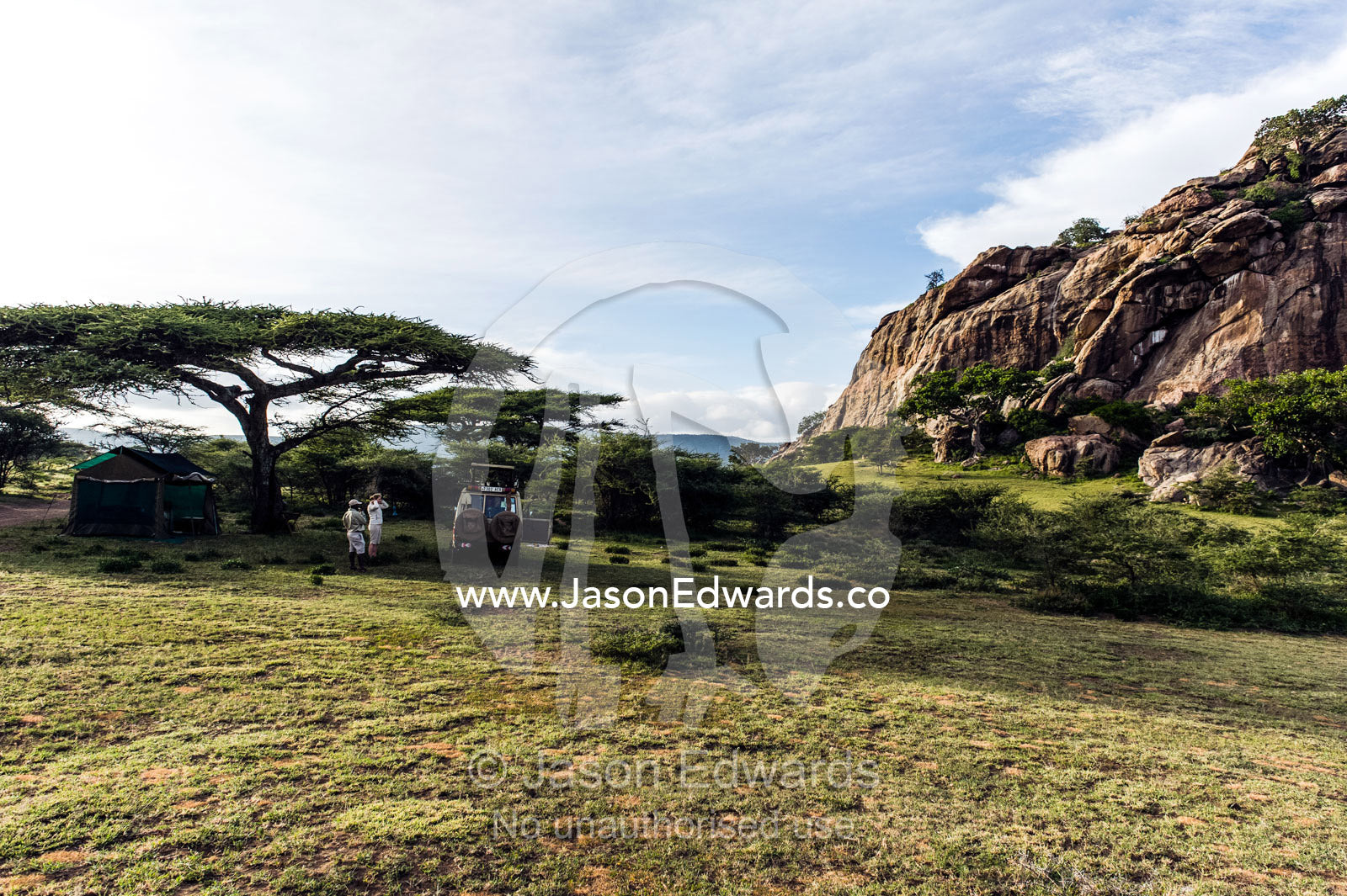 Tourists camping on the savannah at the base of a large rock outcrop. Arusha, Nasera Rock, Angata Kiti Valley, Ngorongoro Conservation Area, Crater Highlands, Tanzania.