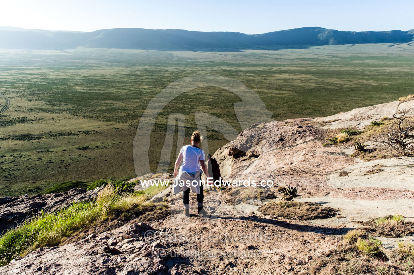 A tourist carefully descends a steep rocky outcrop overlooking a distant savannah plain. Arusha, Nasera Rock, Angata Kiti Valley, Ngorongoro Conservation Area, Crater Highlands, Tanzania.
