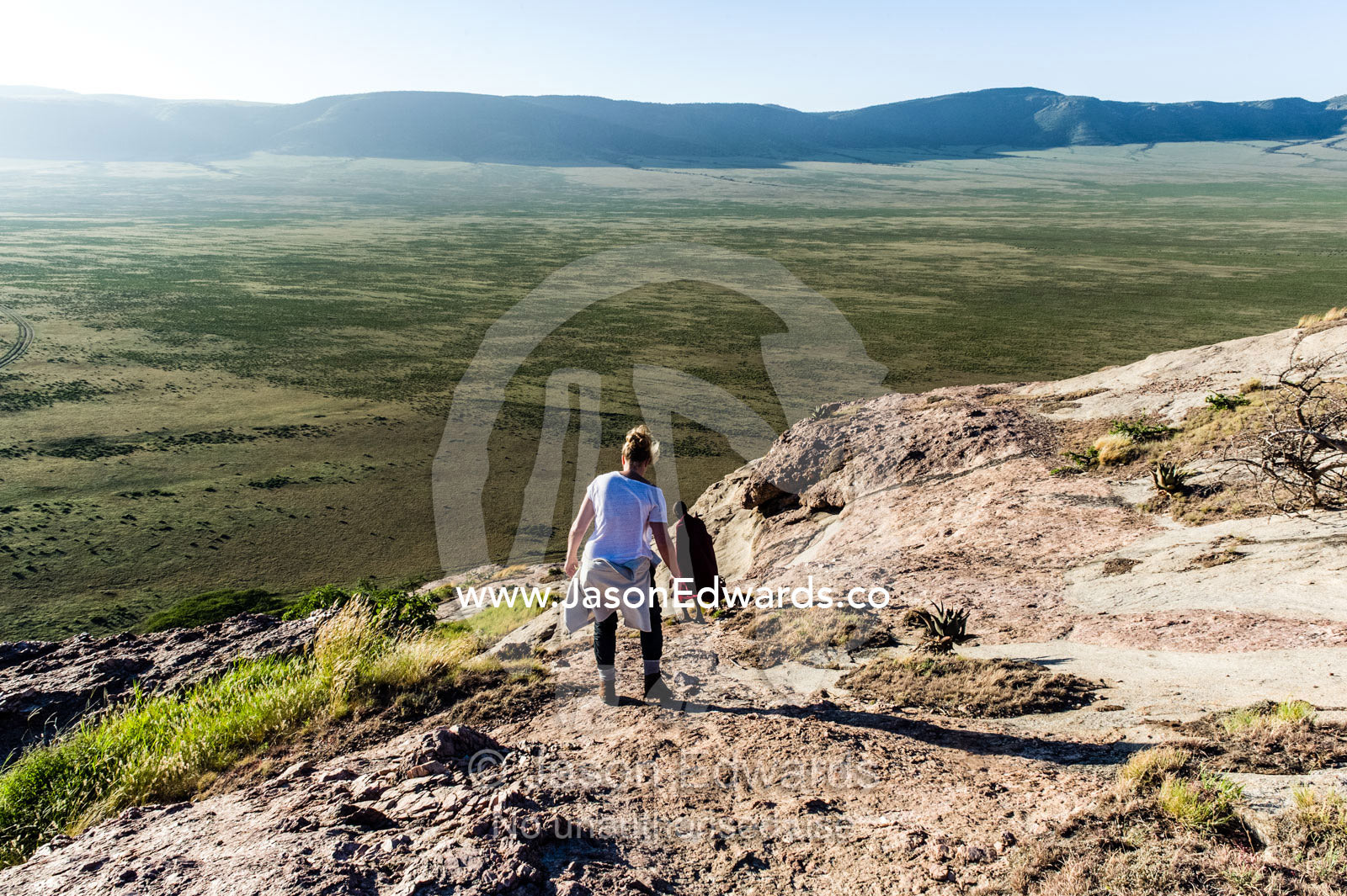 A tourist carefully descends a steep rocky outcrop overlooking a distant savannah plain. Arusha, Nasera Rock, Angata Kiti Valley, Ngorongoro Conservation Area, Crater Highlands, Tanzania.