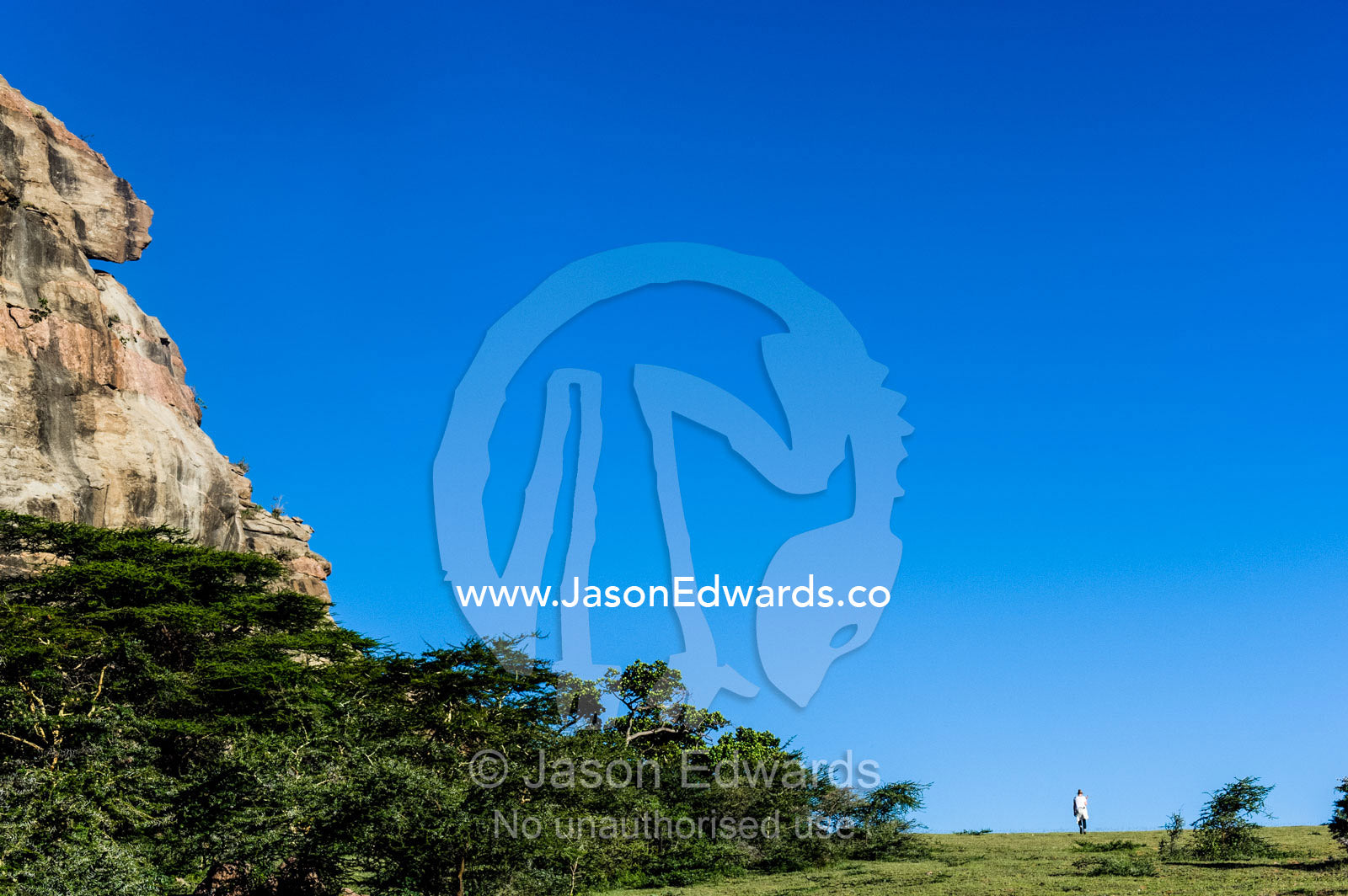 A tourist hiking past an Acacia woodland at the base of a rocky outcrop. Arusha, Nasera Rock, Angata Kiti Valley, Ngorongoro Conservation Area, Crater Highlands, Tanzania.