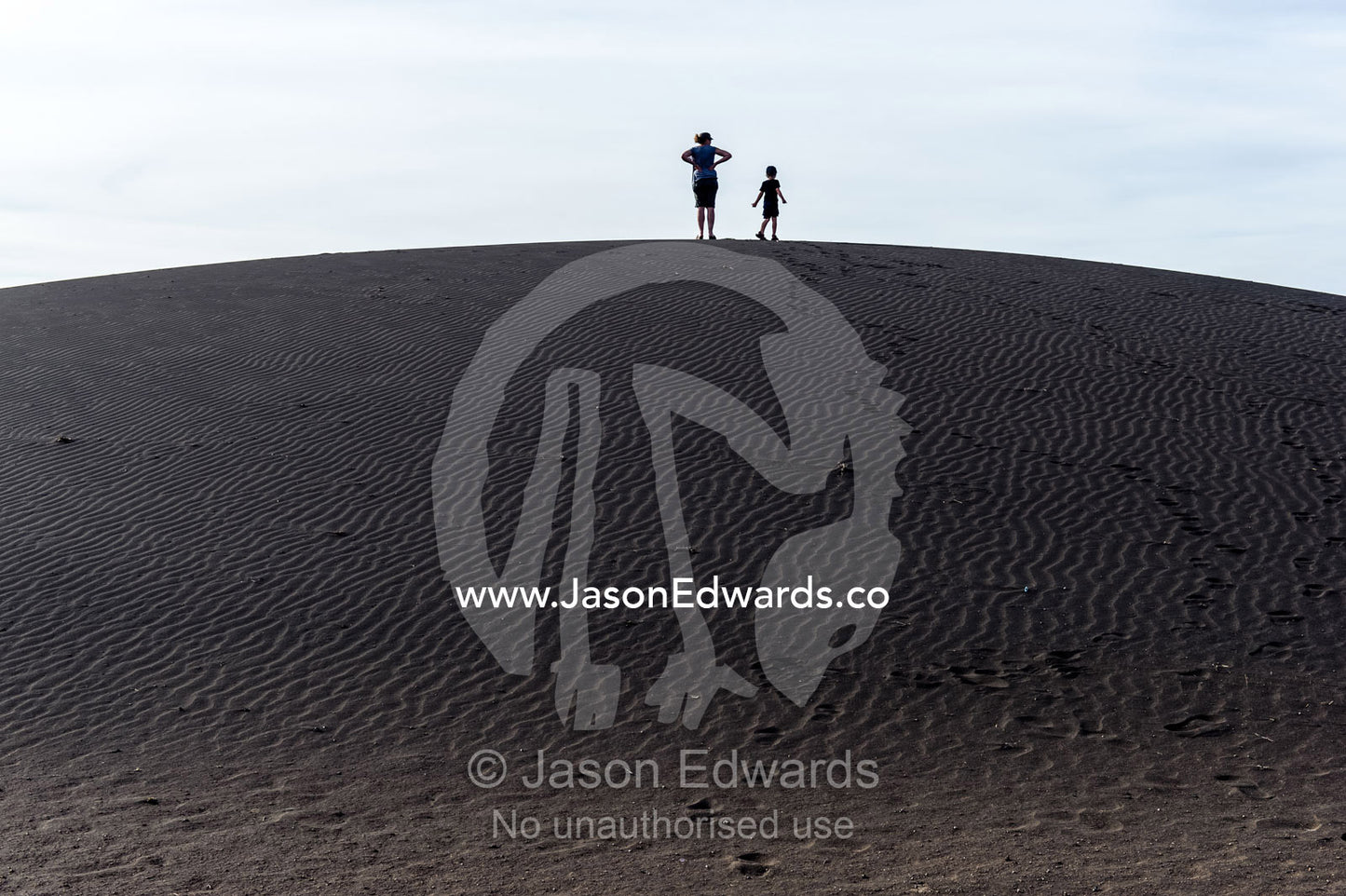 Mother and son tourists on a safari climb to the top of a black volcanic sand dune. Shifting Sands, Ngorongoro Conservation Area, Ngorongoro District, Tanzania.
