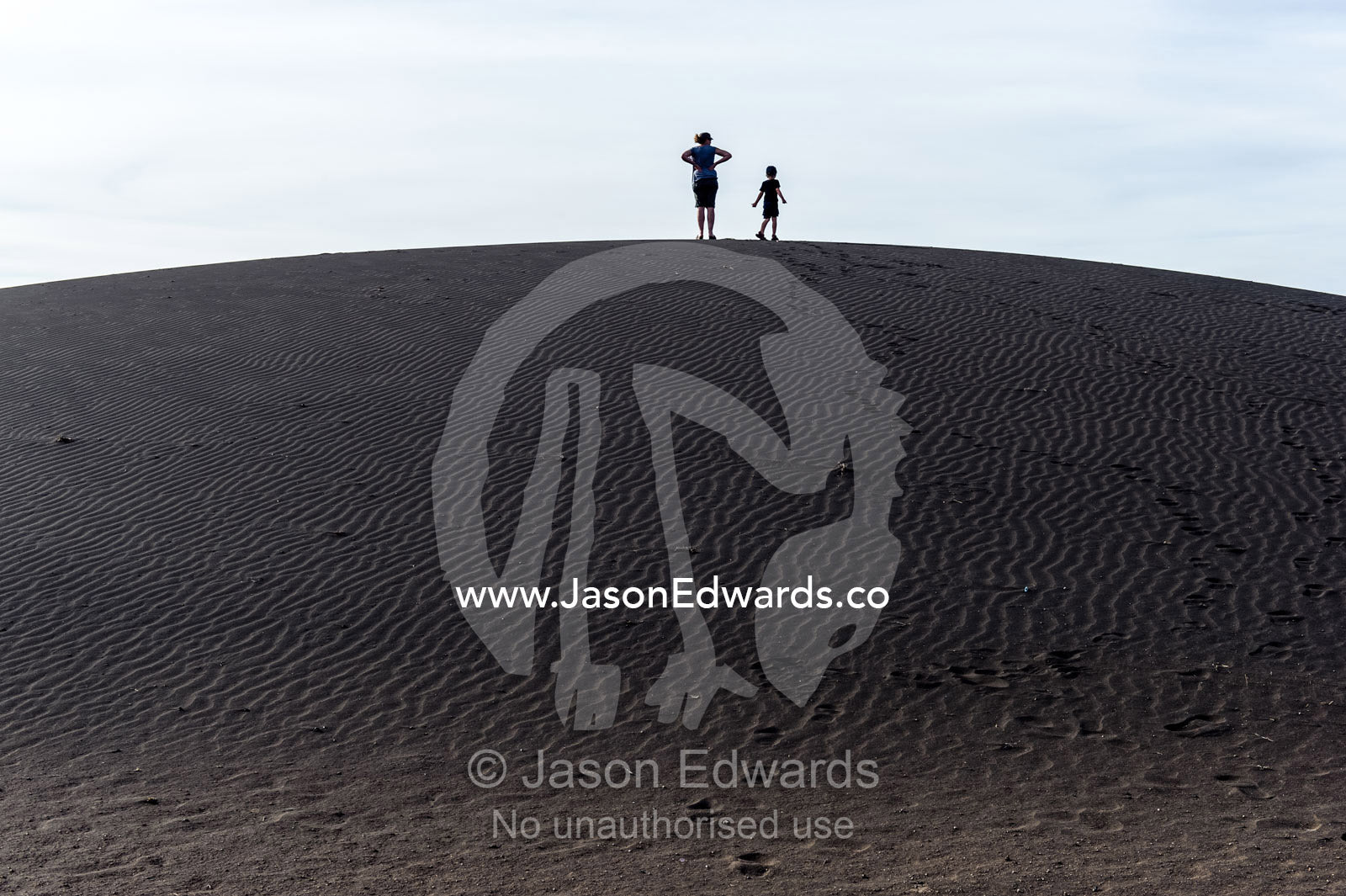 Mother and son tourists on a safari climb to the top of a black volcanic sand dune. Shifting Sands, Ngorongoro Conservation Area, Ngorongoro District, Tanzania.