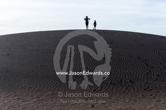 Mother and son tourists on a safari climb to the top of a black volcanic sand dune. Shifting Sands, Ngorongoro Conservation Area, Ngorongoro District, Tanzania.