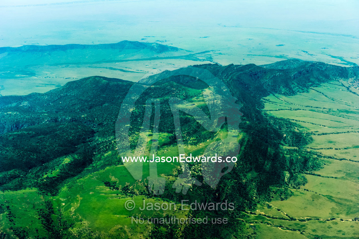 Forested volcanic mountain slopes rise above the vast, green savannah plain. Crater Highlands, Ngorongoro Highlands, Serengeti National Park, Serengeti, Tanzania.