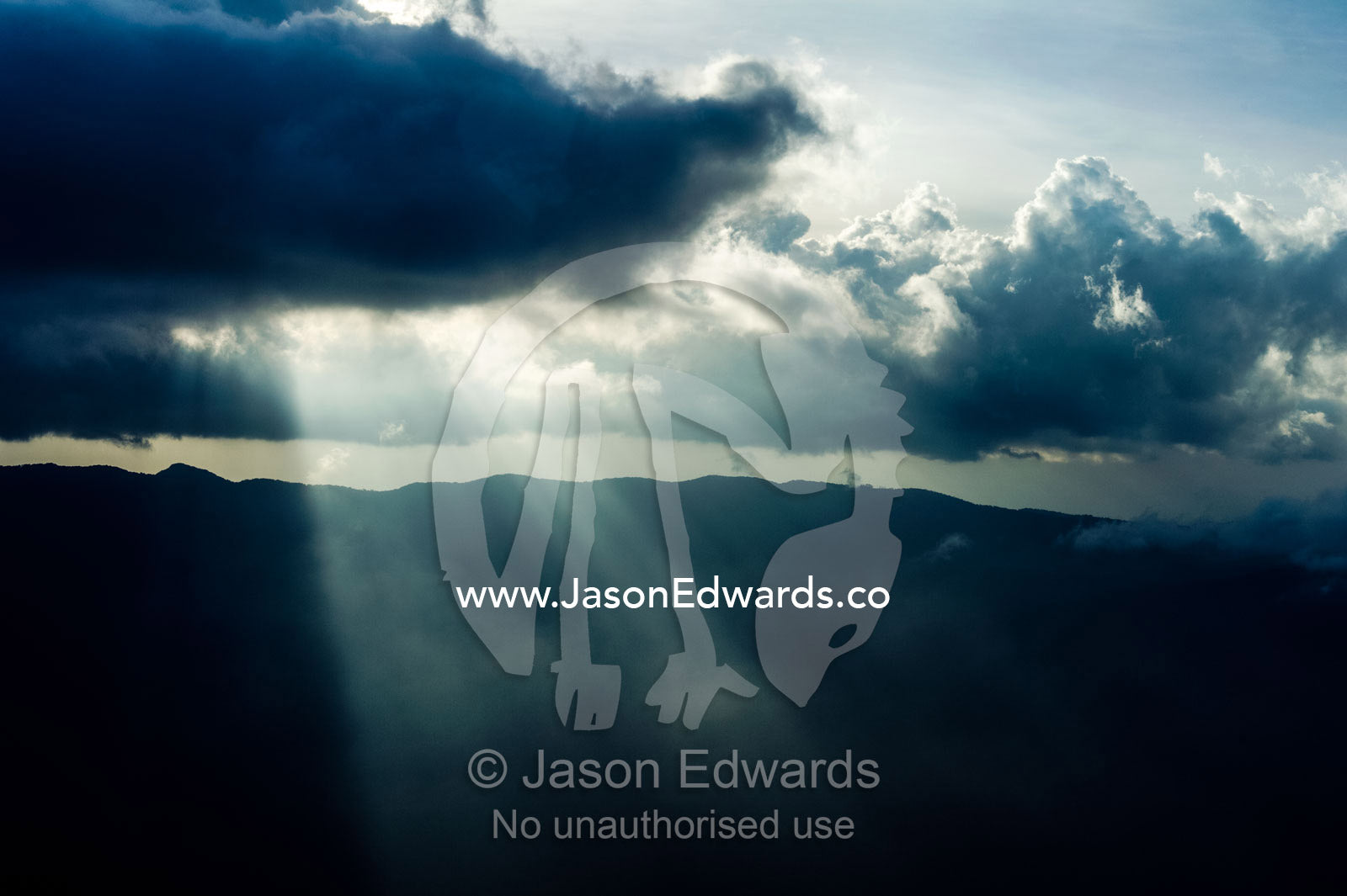 Sun rays pierce mist and cloud descending over the summits of mountain peaks at dusk. Crater Highlands, Arusha District, Tanzania.