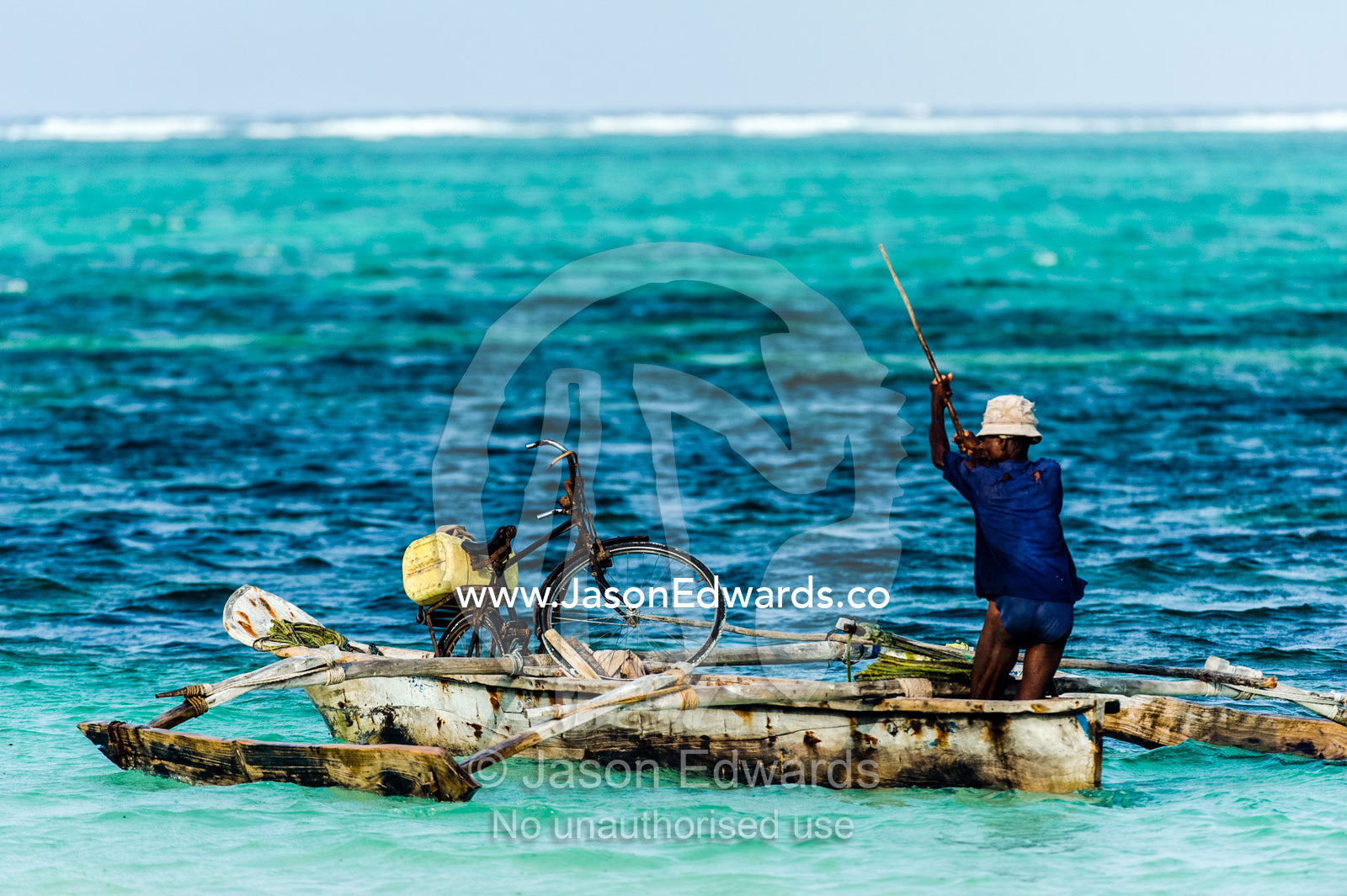 A fisherman poles his wooden trimaran dhow and bicycle through the shallows towards a reef. Bwejuu, Echo Beach Hotel, Echo Beach, Unguja Island, Zanzibar, Tanzania, Africa.