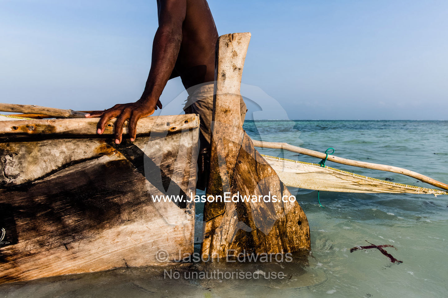 A fisherman moors his wooden trimaran sailing dhow in the shallows at sunset. Bwejuu, Unguja Island, Zanzibar, Tanzania.