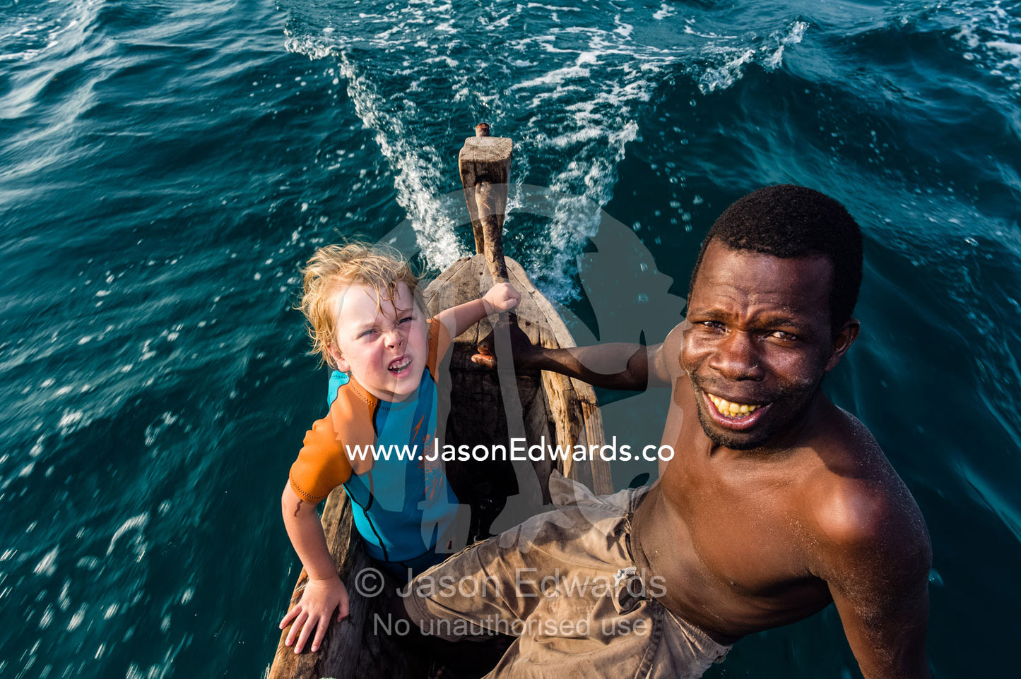 A small boy and a fisherman steer his wooden trimaran sailing dhow in the shallow turquoise seas of the Indian Ocean. Bwejuu, Unguja Island, Zanzibar, Tanzania.