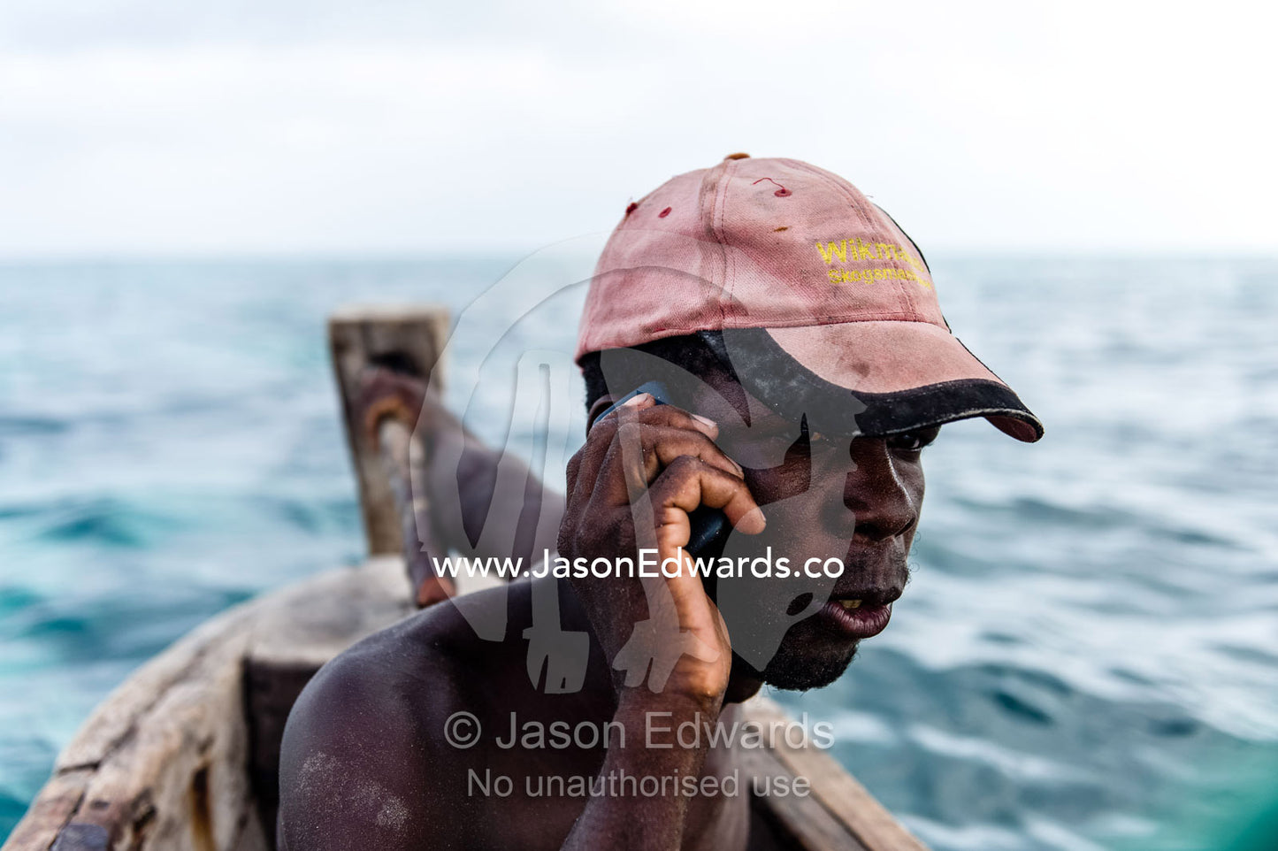 A fisherman on his mobile phone, steers his wooden trimaran sailing dhow in the Indian Ocean. Bwejuu, Unguja Island, Zanzibar, Tanzania, Africa.