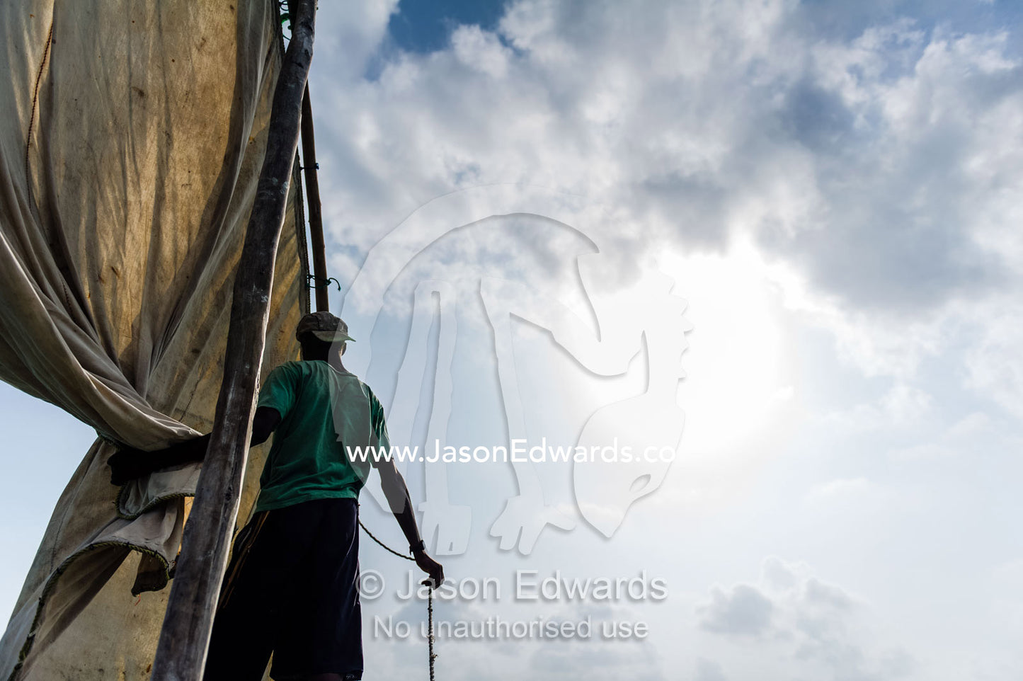A deckhand on a wooden trimaran dhow working a tarpaulin sail under a blistering sun. Bwejuu, Unguja Island, Zanzibar, Tanzania, Africa.
